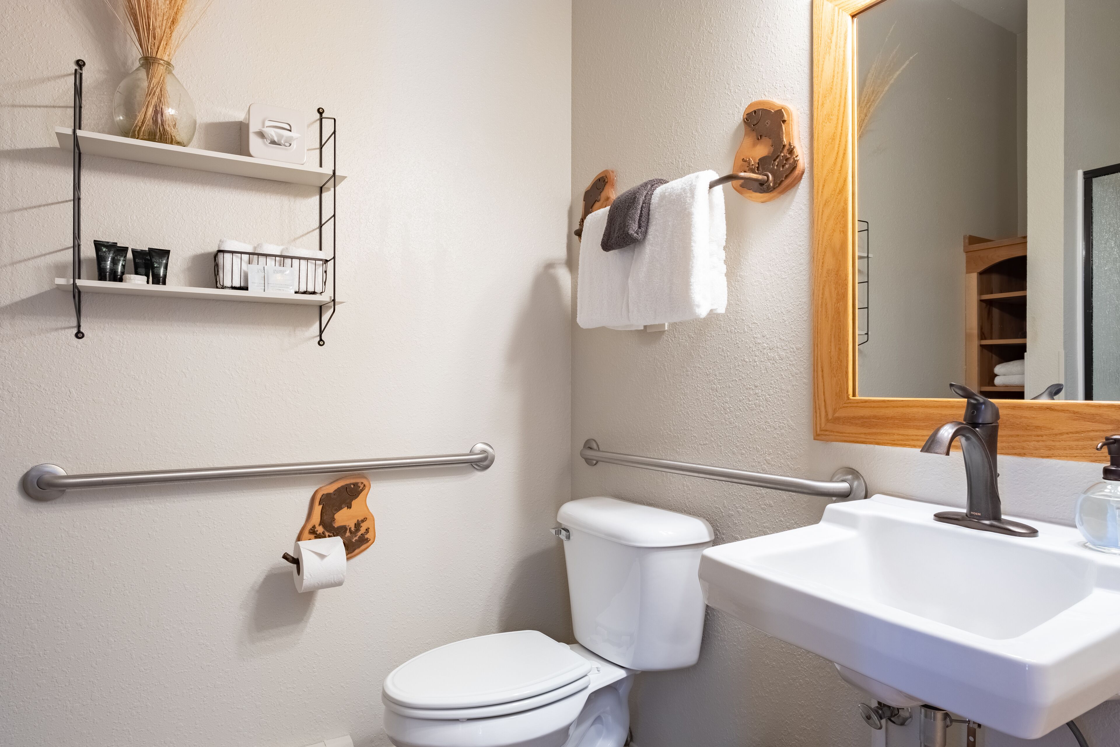 Un baño con inodoro blanco, un lavabo y un espejo. Hay repisas con artículos de aseo y un perchero para toallas.