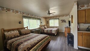 Un dormitorio con dos camas, ventilador de techo, ventana con cortinas, televisión montada en la pared y una pequeña kitchenette.