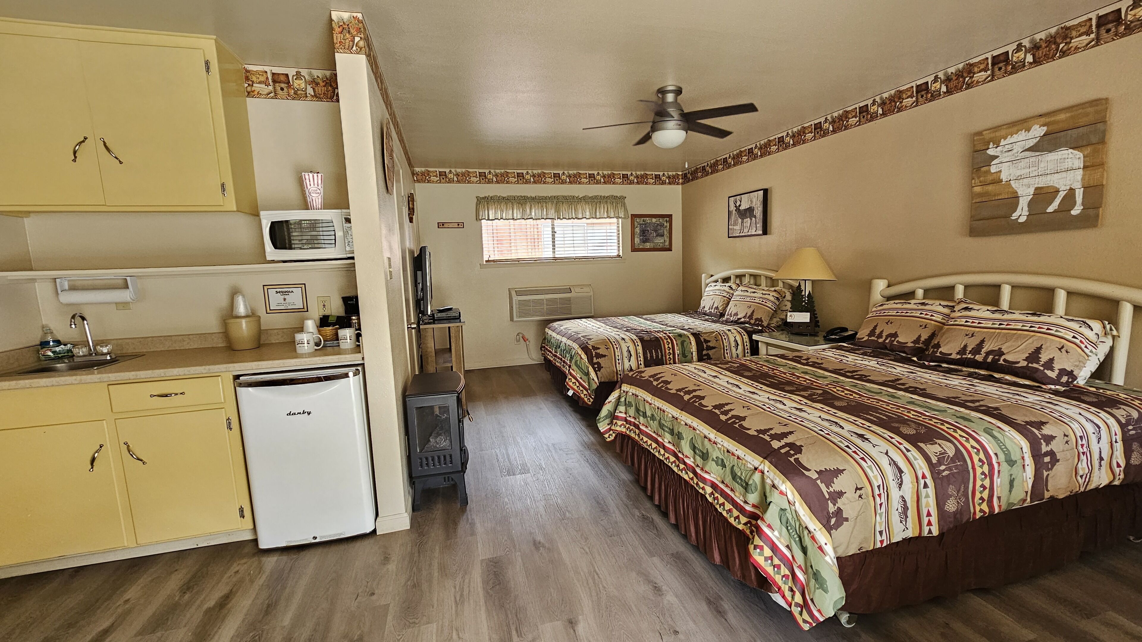A compact room with a kitchenette, two beds with patterned bedding, a ceiling fan, and a window with blinds.