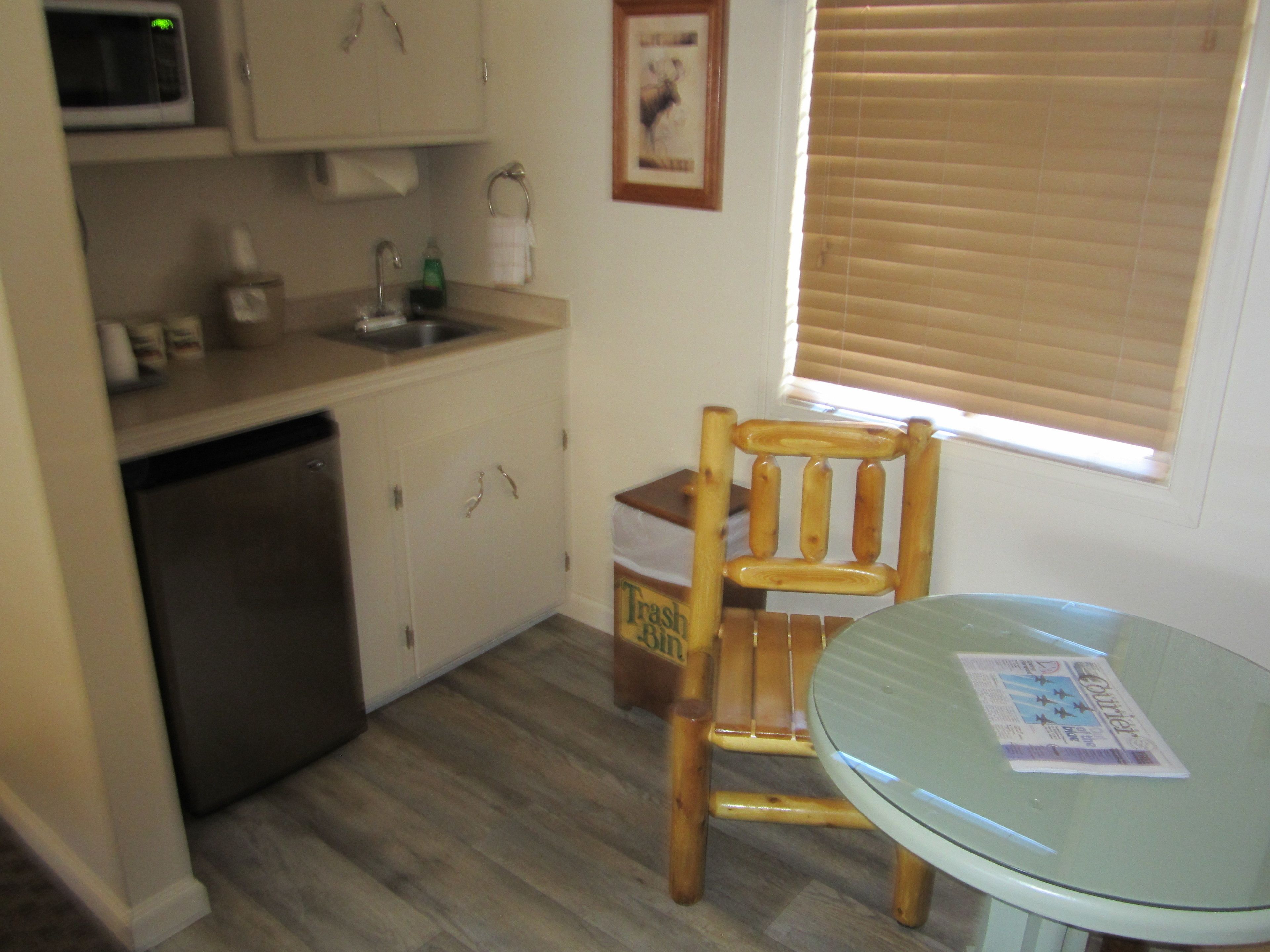 A compact kitchen with a dishwasher, a microwave, a sink, and a wooden chair. There is a window with blinds and a framed picture on the wall.