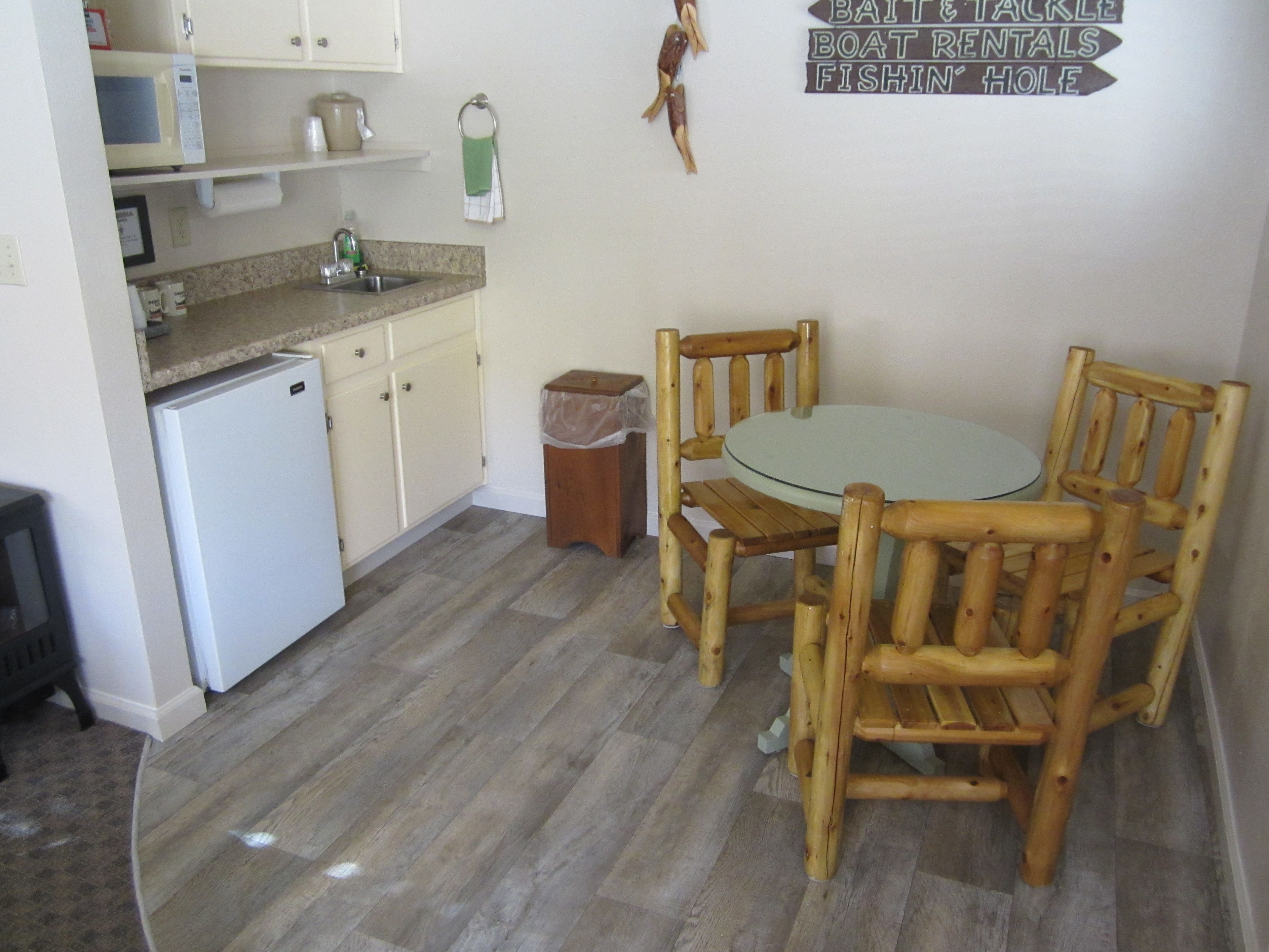 Una cocina compacta equipada con refrigerador, microondas y lavamanos. Hay una mesa de madera con sillas y un cubo de basura. Un cartel en la pared indica atracciones cercanas.
