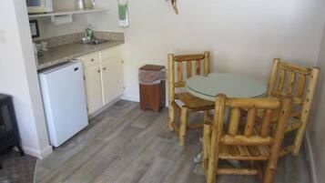 Una cocina compacta equipada con refrigerador, microondas y lavamanos. Hay una mesa de madera con sillas y un cubo de basura. Un cartel en la pared indica atracciones cercanas.