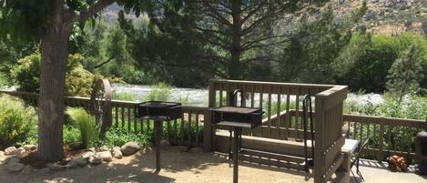 Een terras met barbecuegrills met uitzicht op een rivier, omringd door bomen en een bergachtige achtergrond.
