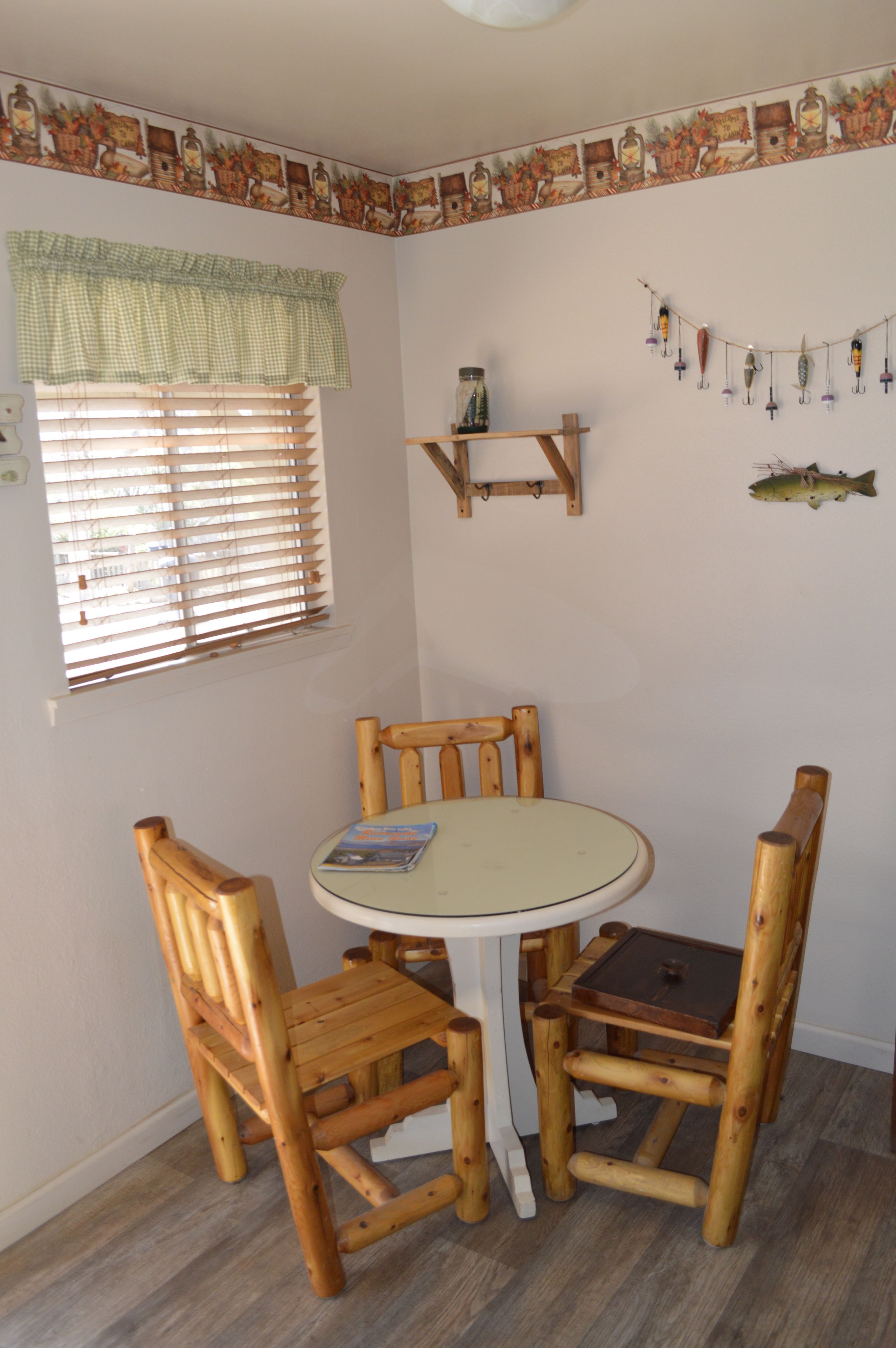 A room with a round table and four wooden chairs, a window with blinds, a shelf with fishing lures, and a wall-mounted fish decoration.