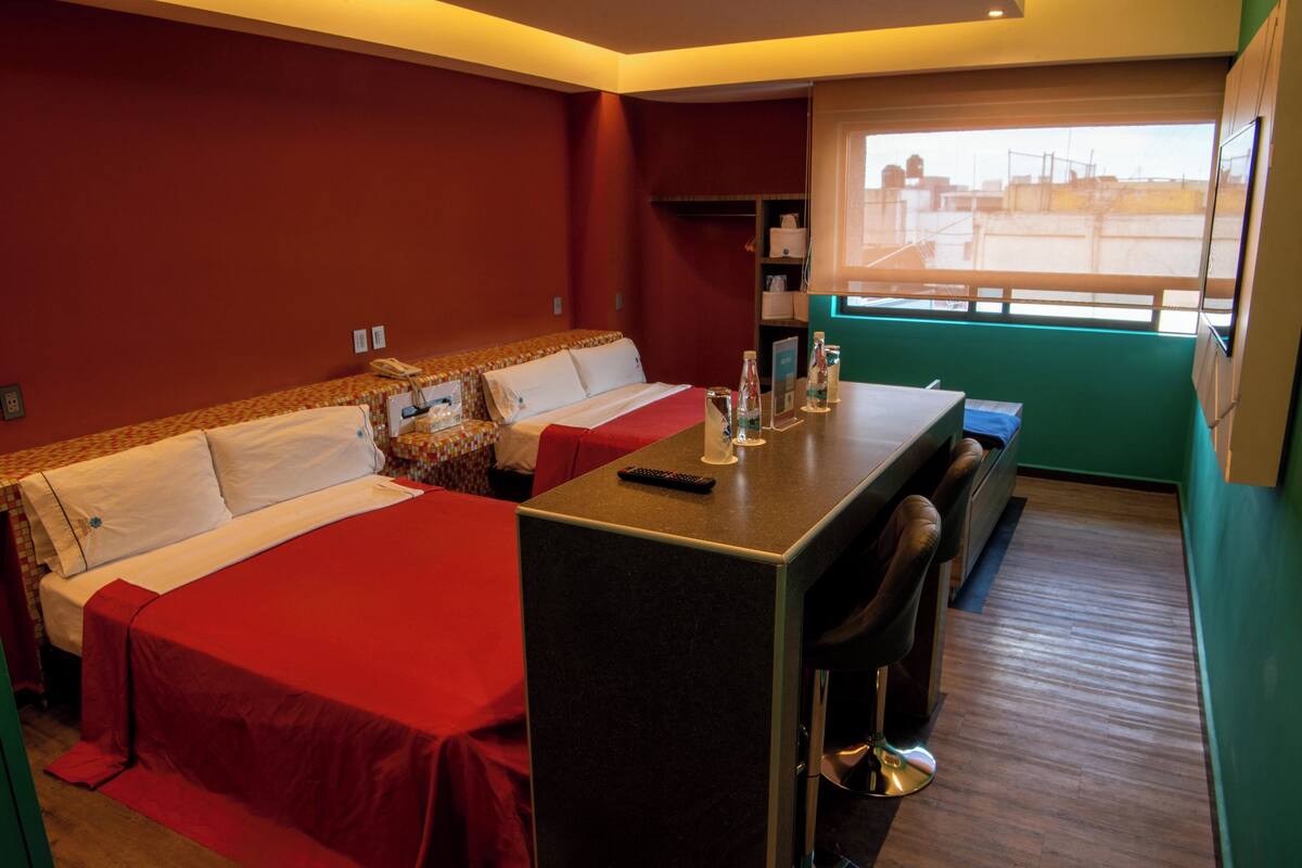 Ein Hotelzimmer mit zwei Betten, einem kleinen Tisch, einem Barhocker und einem Fenster mit Blick auf Gebäude.