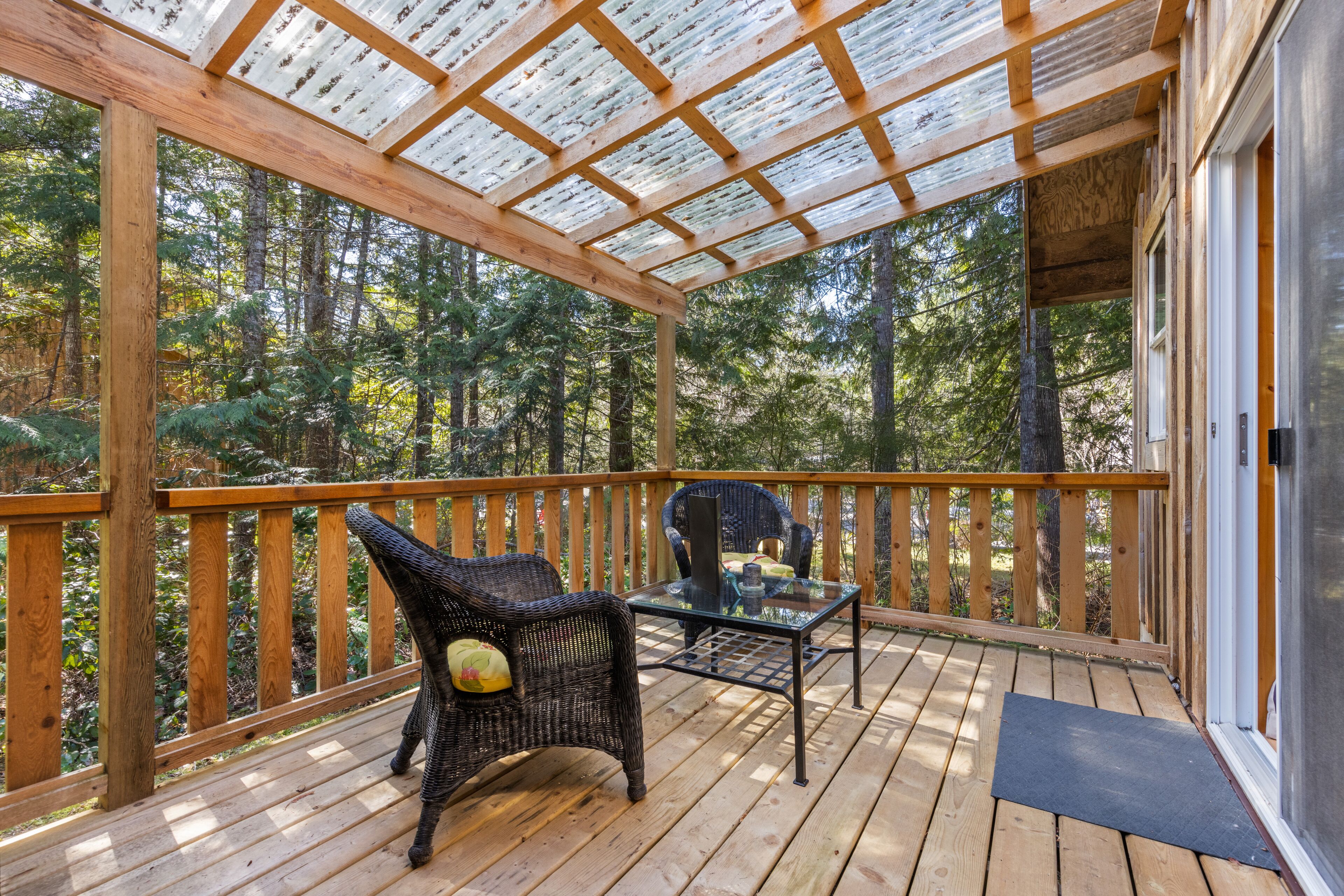 A wooden deck with a glass table and wicker chairs, surrounded by a forest.