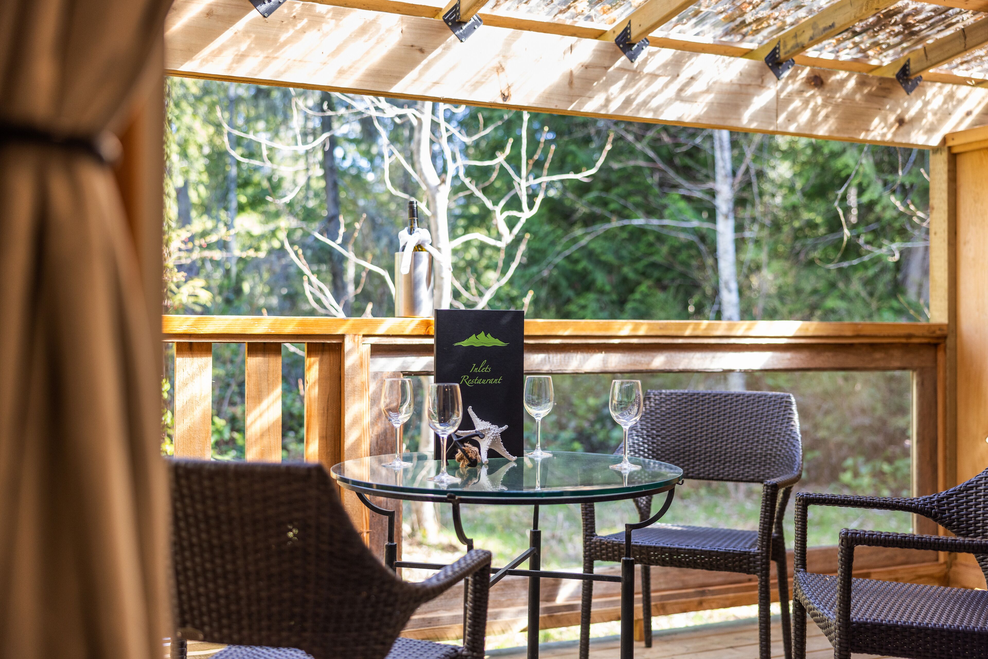 A glass table with four chairs and a menu stand in a wooden deck area with a view of trees.