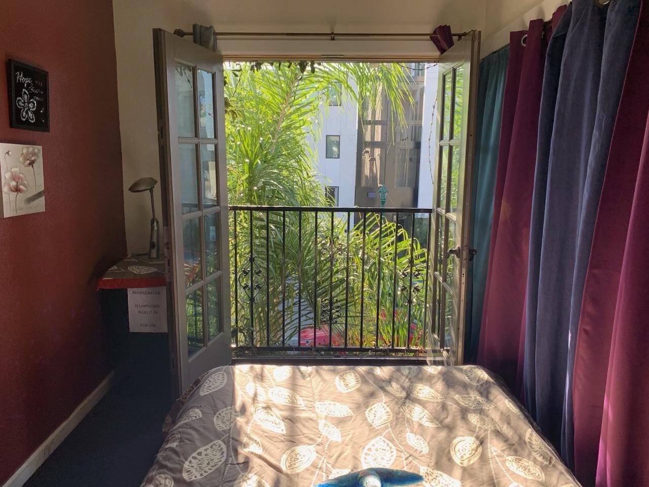 Una habitación con cama, ventana con vista a vegetación, refrigerador y un cuadro en la pared.