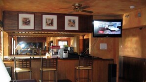 Una zona de bar con mobiliario de madera, un televisor montado en la pared y cuadros enmarcados sobre la barra.