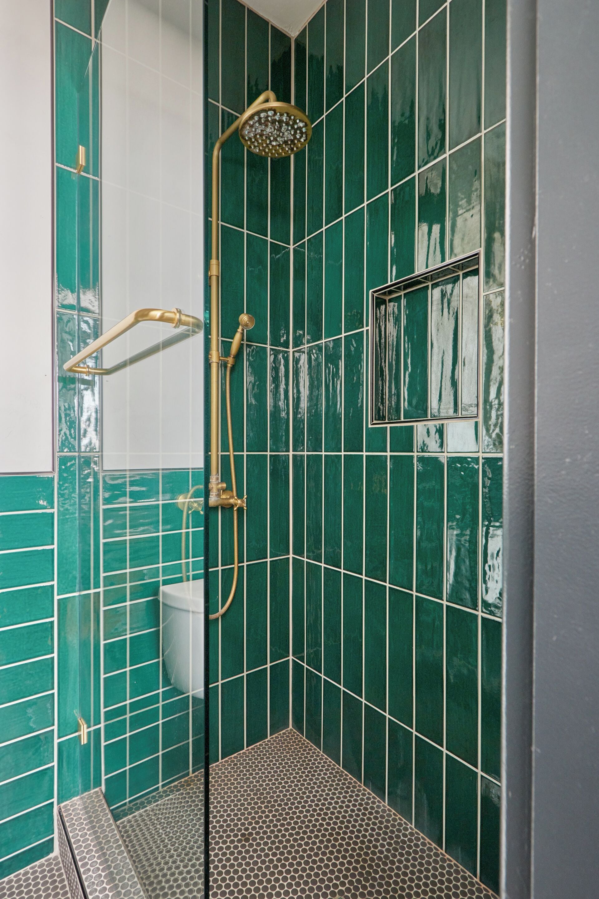 Un baño con paredes de azulejos verdes, ducha con ducha dorada y un inodoro.