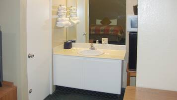 Baño con lavamanos, espejo, barra para toallas y una cama visible en el reflejo.