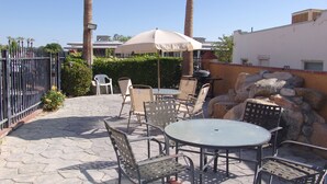 Un patio al aire libre con suelo de piedra, sillas de metal, una mesa redonda y una sombrilla blanca.