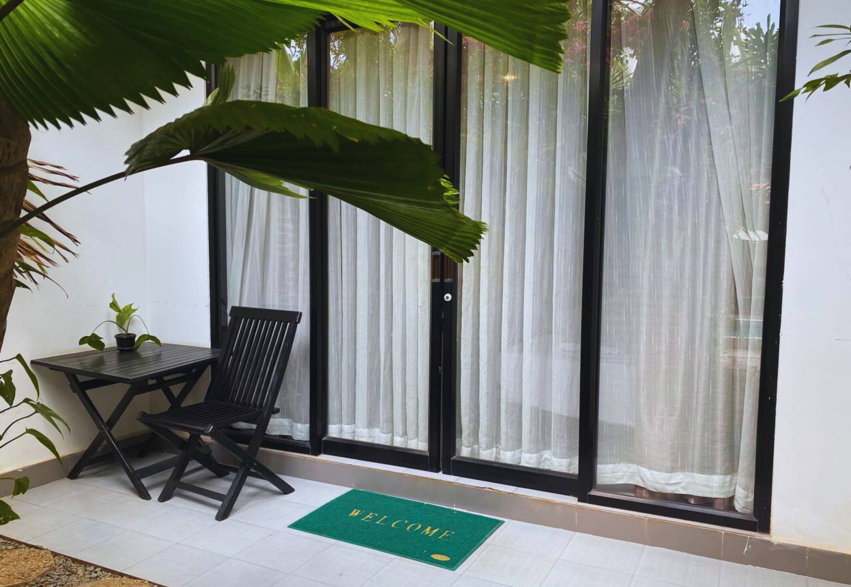 A room with a black table and chair, a green welcome mat, and large windows with sheer curtains.