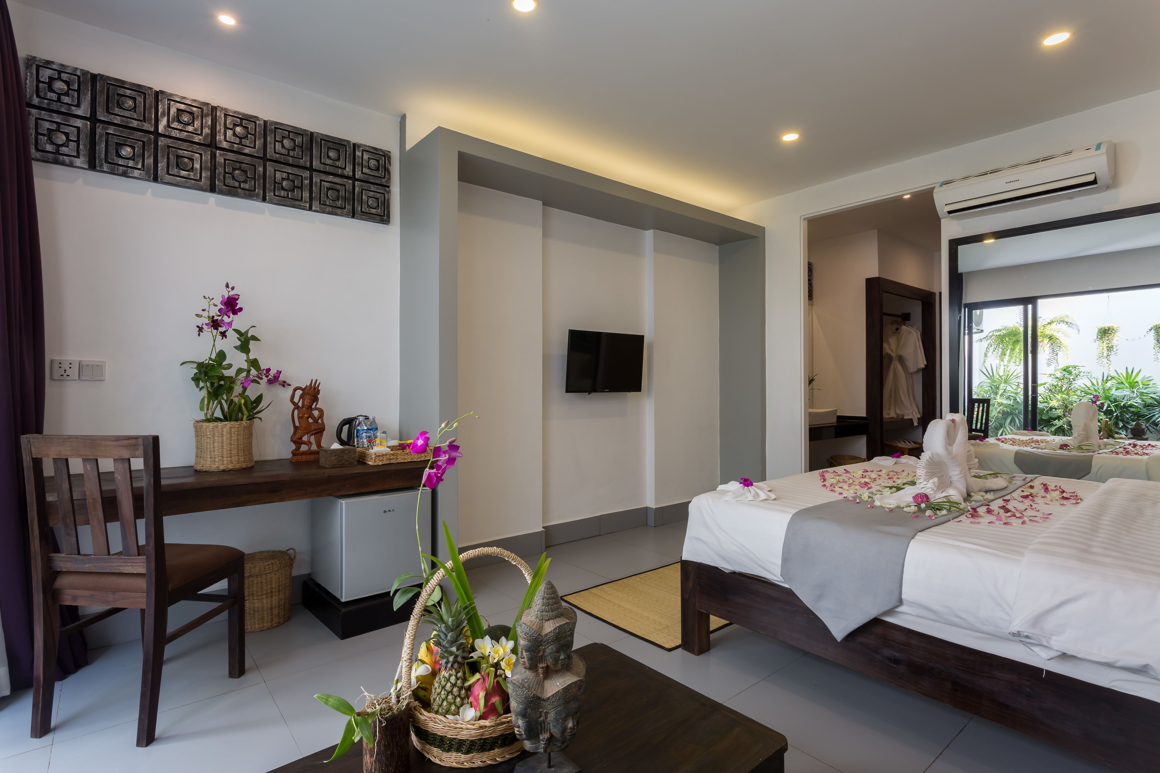 Une chambre d’hôtel moderne, équipée d’un lit, d’un bureau et d’une chaise. Un téléviseur est fixé au mur, une fenêtre offre une vue sur la verdure, et un élément décoratif orne le mur au-dessus du bureau.