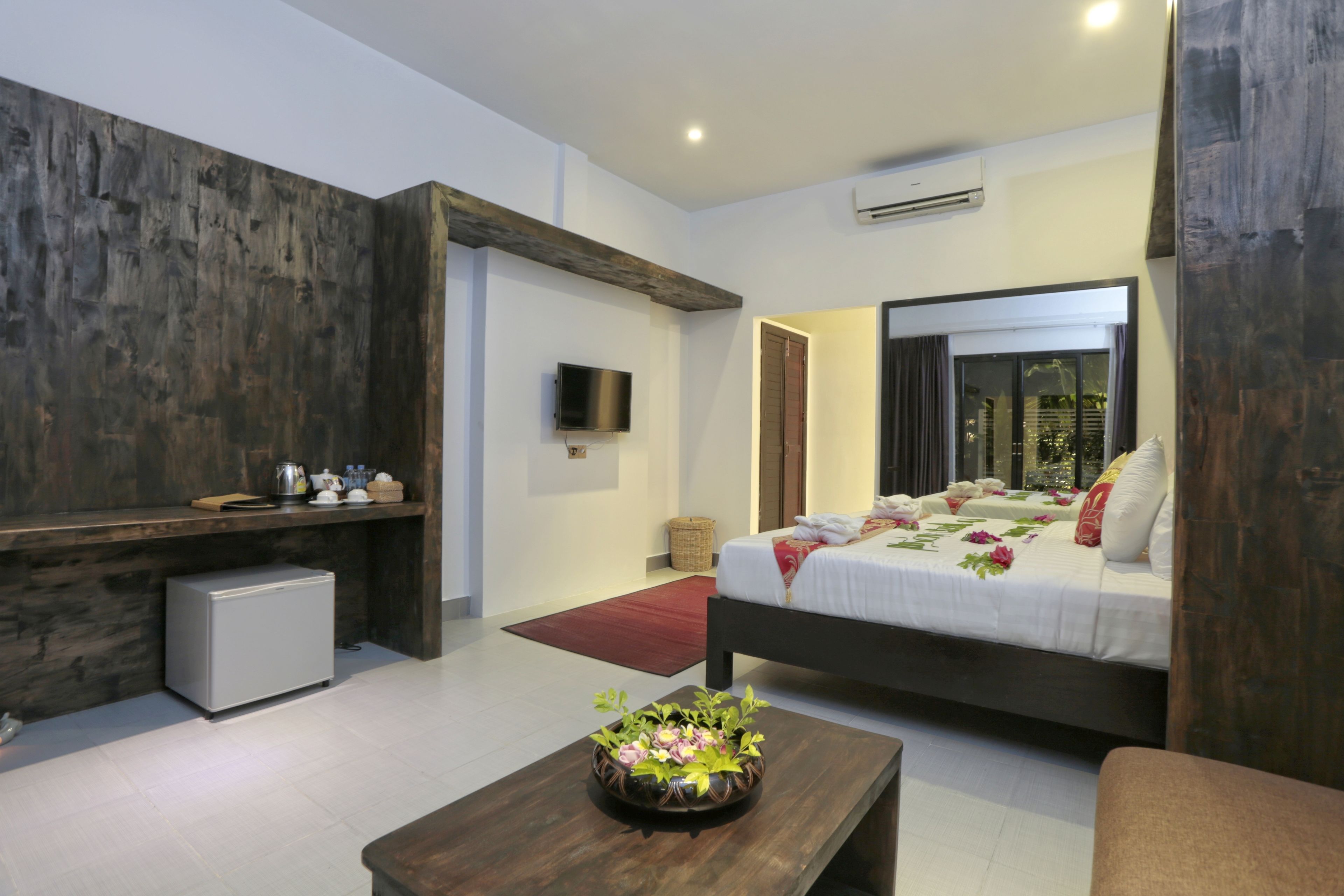 A modern bedroom with a large bed, a small bedside table, a TV mounted on the wall, and a compact refrigerator.