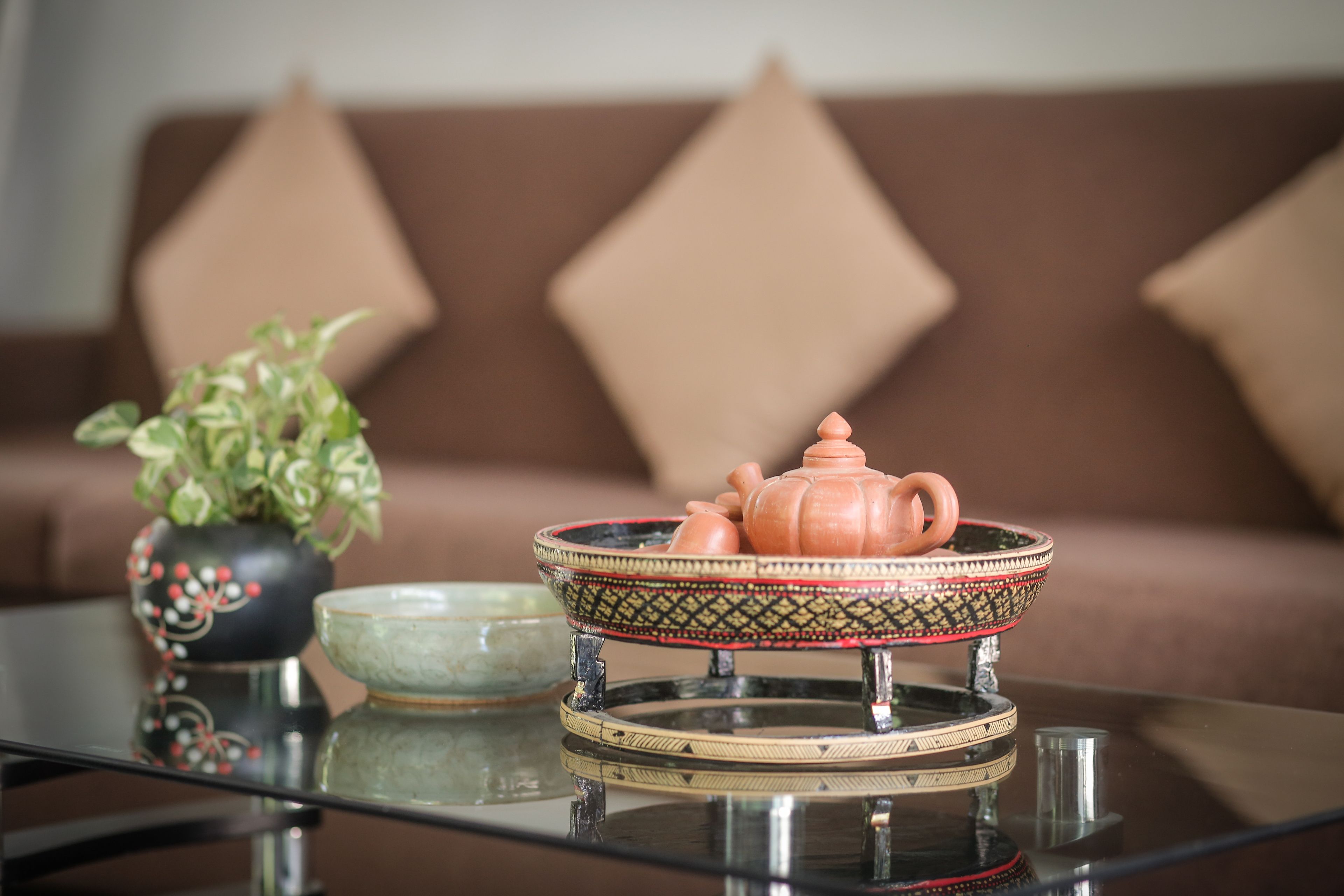 A table with a teapot and a bowl on a glass surface, a potted plant, and a brown sofa with diamond-shaped cushions in the background.