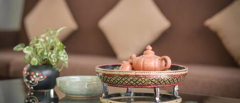 A table with a teapot and a bowl on a glass surface, a potted plant, and a brown sofa with diamond-shaped cushions in the background.