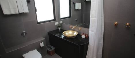 Un baño moderno con tocador negro, lavamanos dorado, inodoro blanco y un espejo grande.
