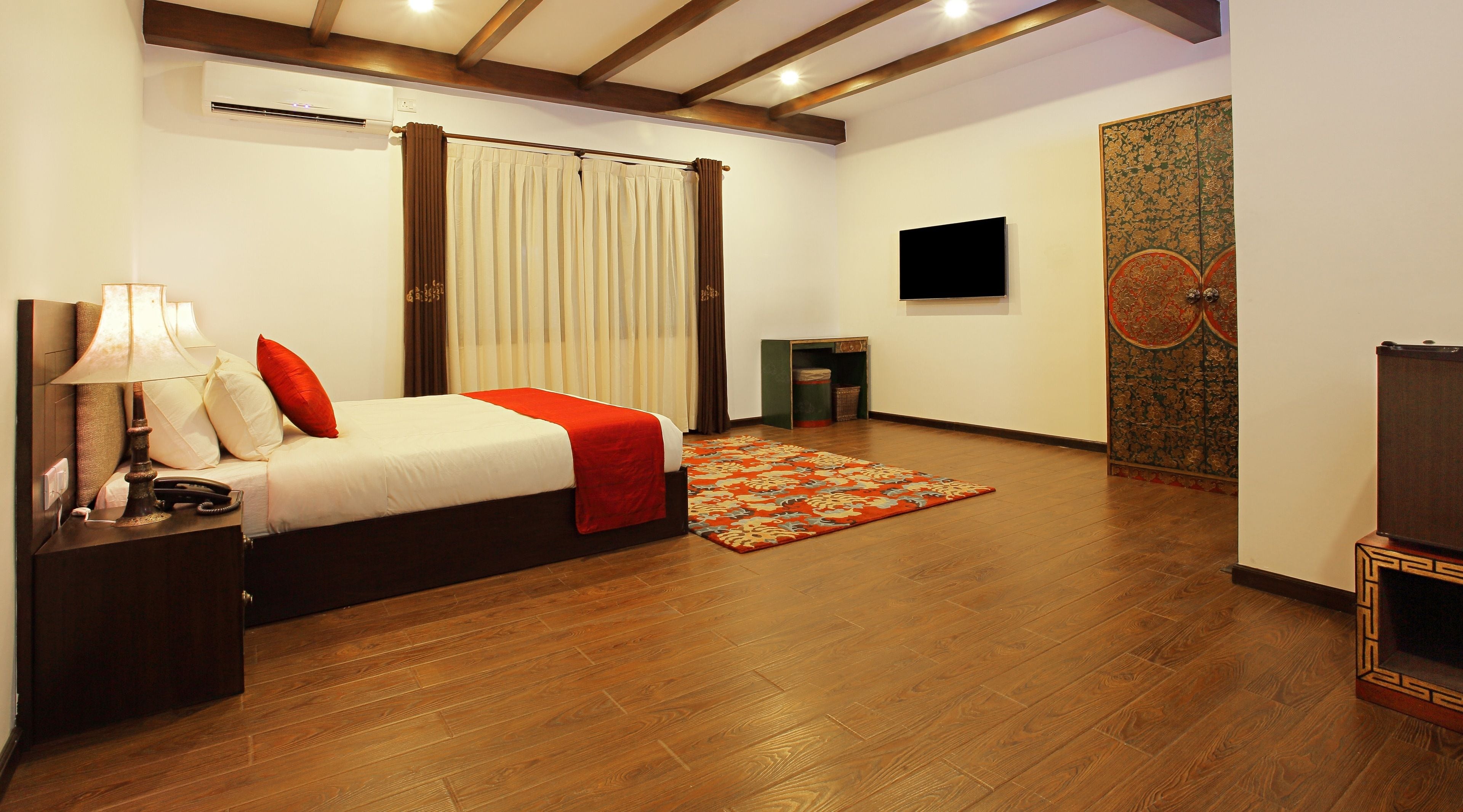 A hotel room with a bed, a nightstand, a lamp, a TV, a small table, and a patterned rug.