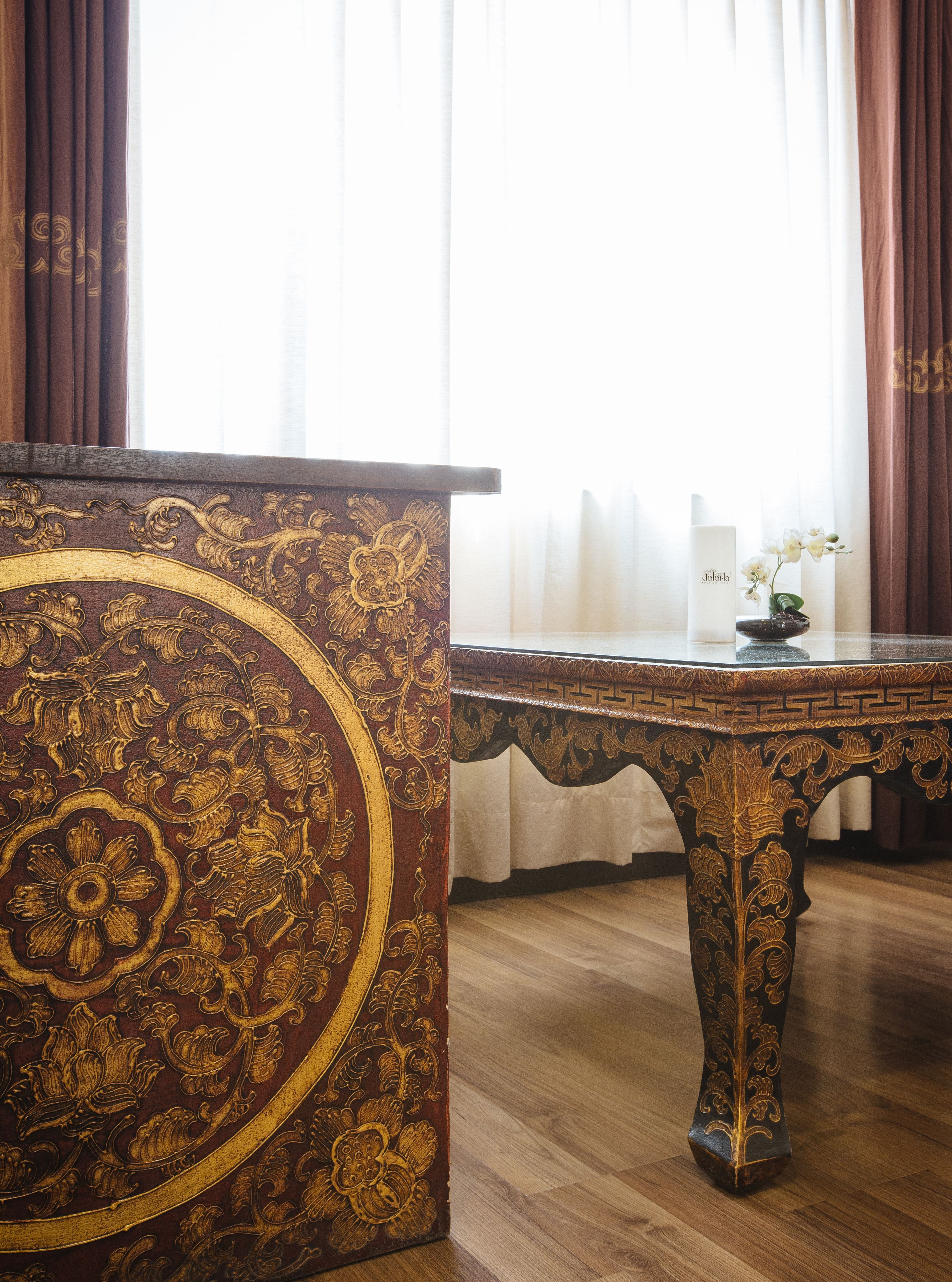 Una habitación con piso de madera, una mesa marrón oscuro y ornamentada con detalles dorados, y un florero blanco con flores sobre una mesa a juego.