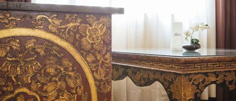 Una habitación con piso de madera, una mesa marrón oscuro y ornamentada con detalles dorados, y un florero blanco con flores sobre una mesa a juego.