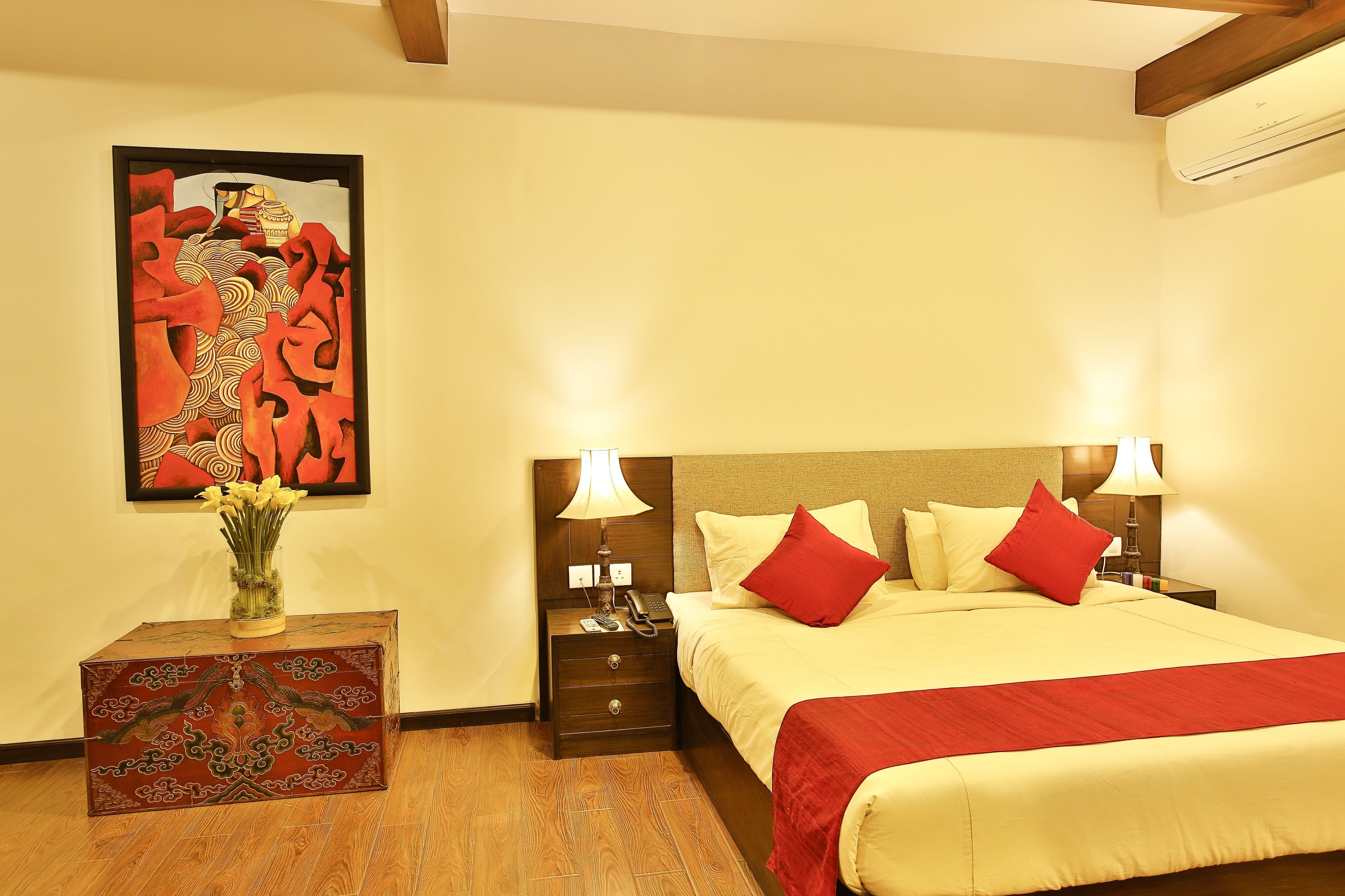 A hotel room with a bed, bedside lamps, a nightstand, a vase with flowers, and a painting on the wall.