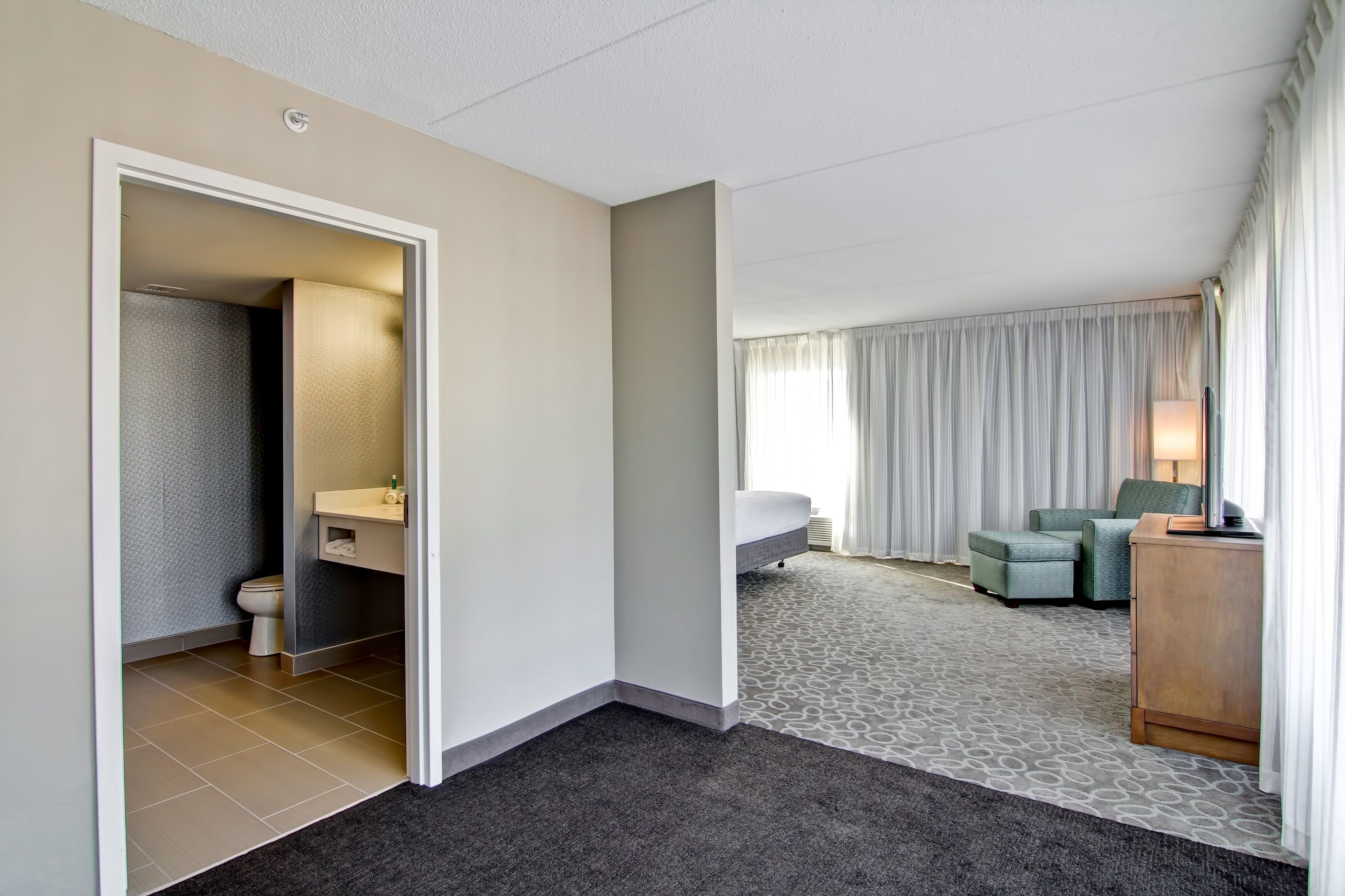 A hotel room with a bathroom, a sitting area with a sofa and armchair, and a desk with a lamp.