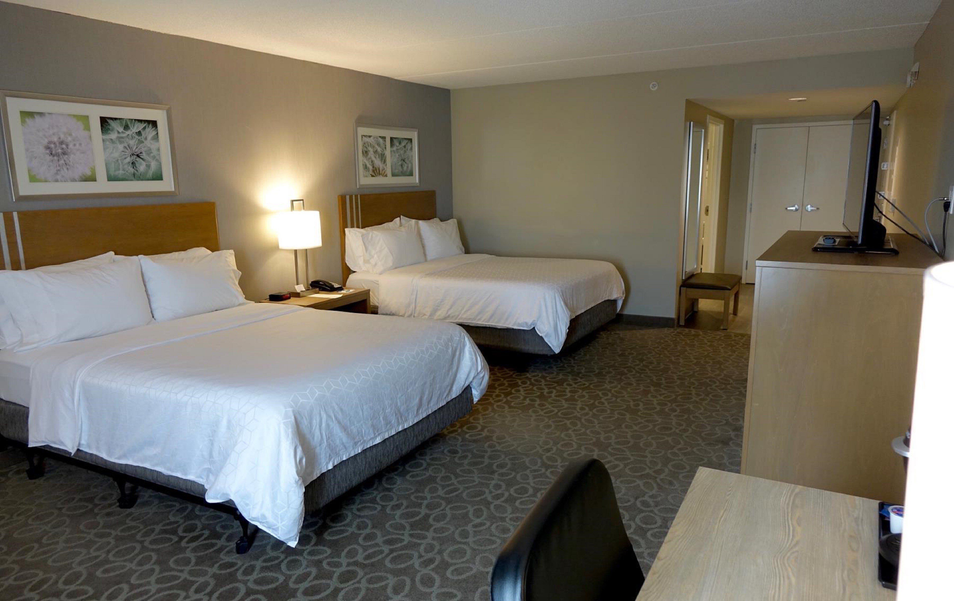 A hotel room with two beds, a desk, a chair, a television, and artwork on the walls.
