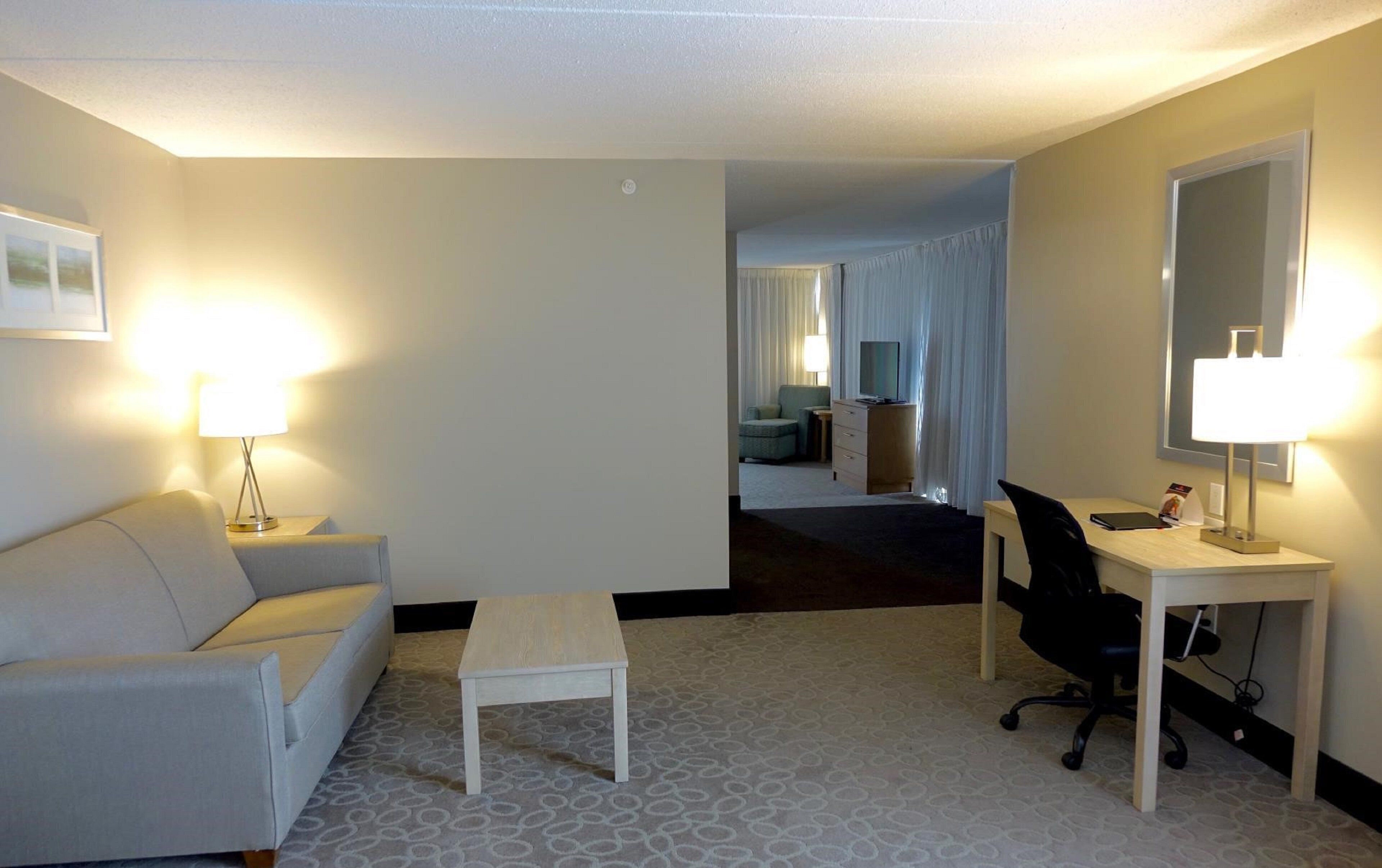 A hotel room with a sofa, a small table, a desk with a chair, and a television.