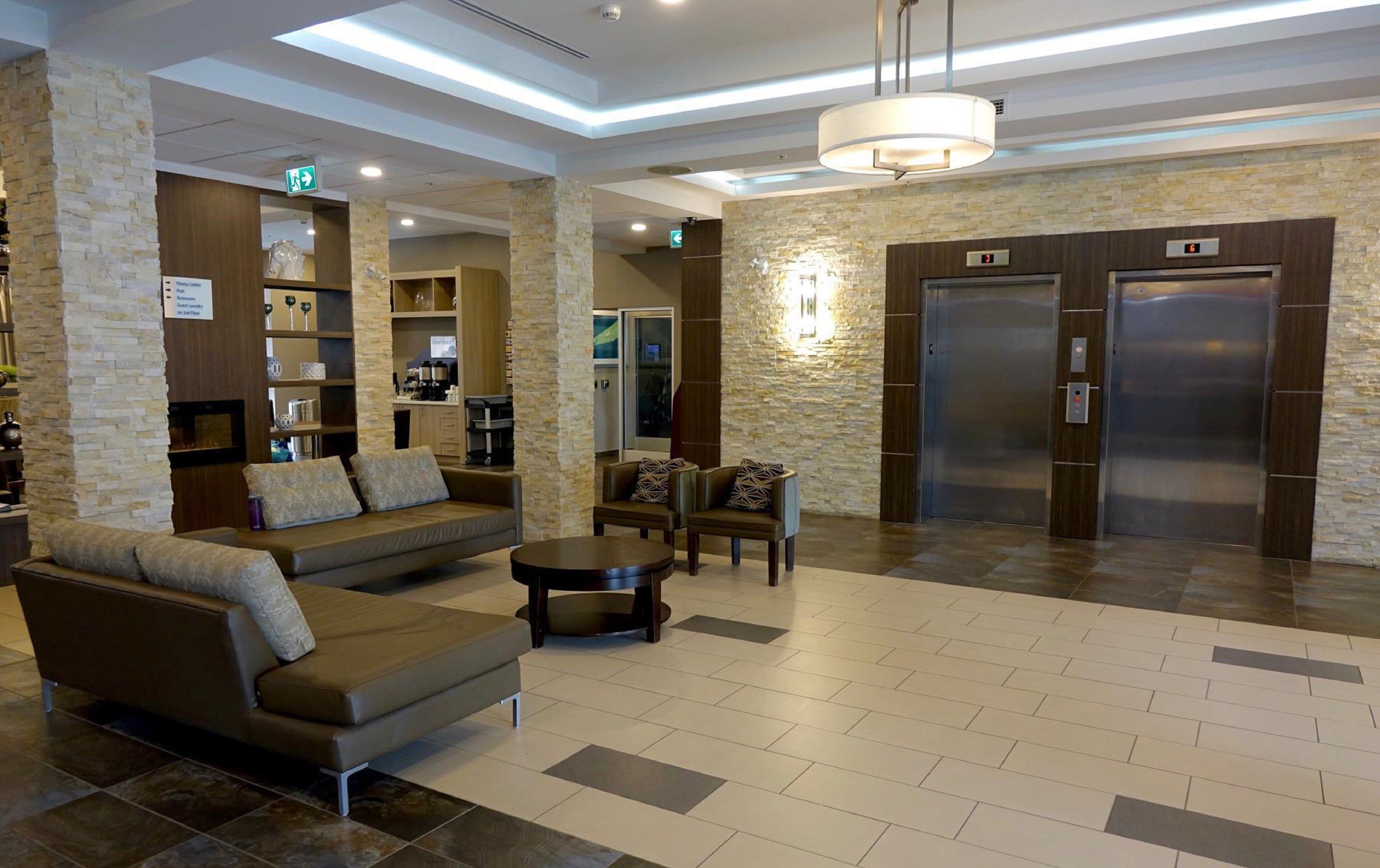 A hotel lobby with a sofa, armchairs, a coffee table, a fireplace, a kitchen area, and two elevators.