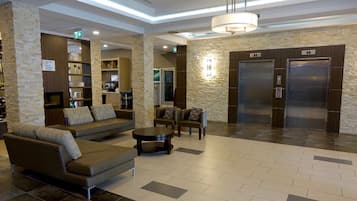 A hotel lobby with a sofa, armchairs, a coffee table, a fireplace, a kitchen area, and two elevators.