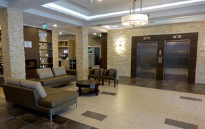 A hotel lobby with a sofa, armchairs, a coffee table, a fireplace, a kitchen area, and two elevators. - Holiday Inn Express & Suites Oshawa Downtown - Toronto Area by IHG (Oshawa)