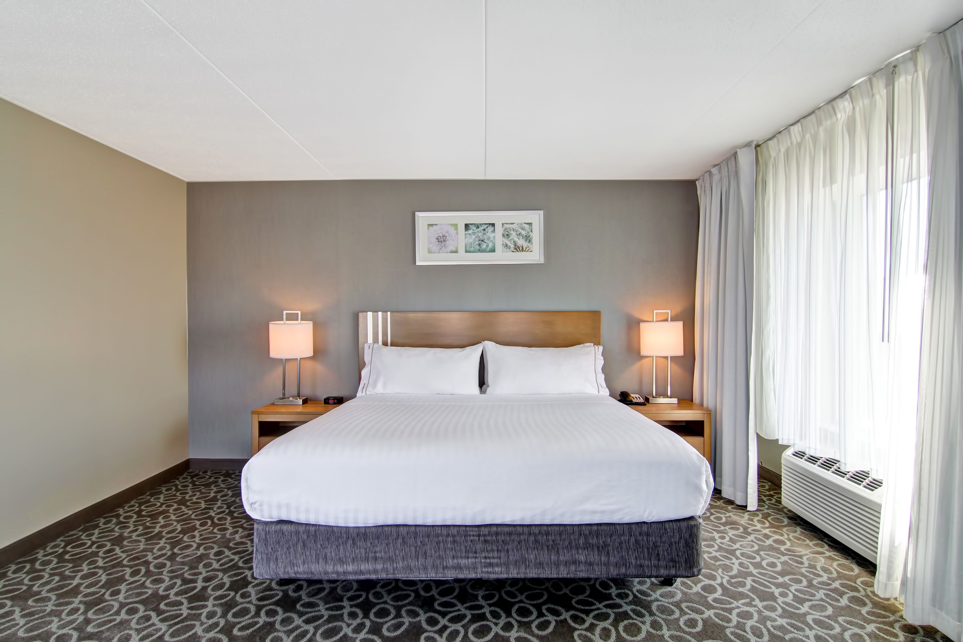 A hotel room with a large bed, two bedside lamps, a framed artwork above the bed, and a window with curtains.