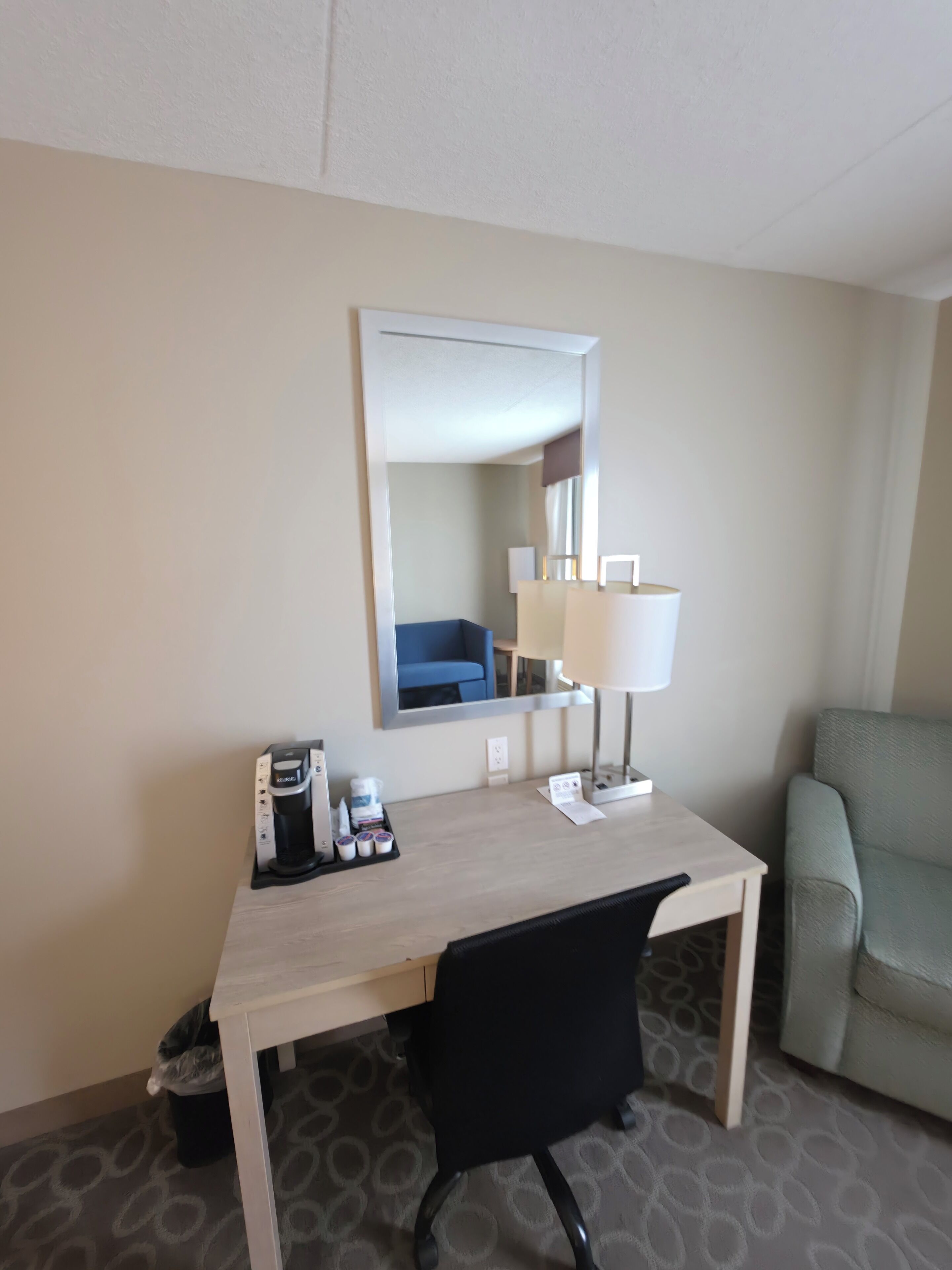 A hotel room with a desk, a chair, a lamp, a coffee maker, and a trash bin.