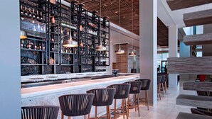 A modern bar area with a long counter, black chairs, and shelves stocked with bottles and glasses.