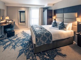 A hotel room with a large bed, a desk, a chair, and a window with curtains.