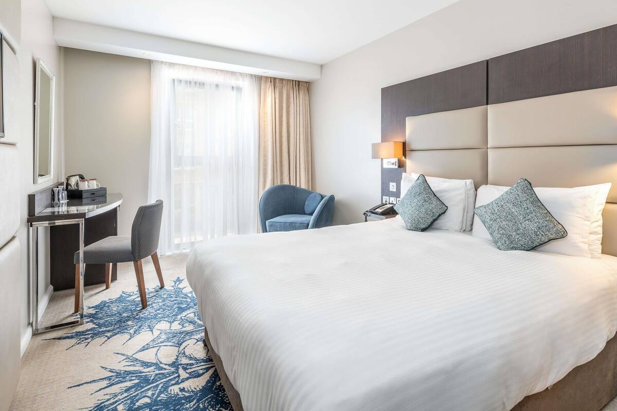 A hotel room with a large bed, a desk with a chair, a lamp, and a window with curtains.