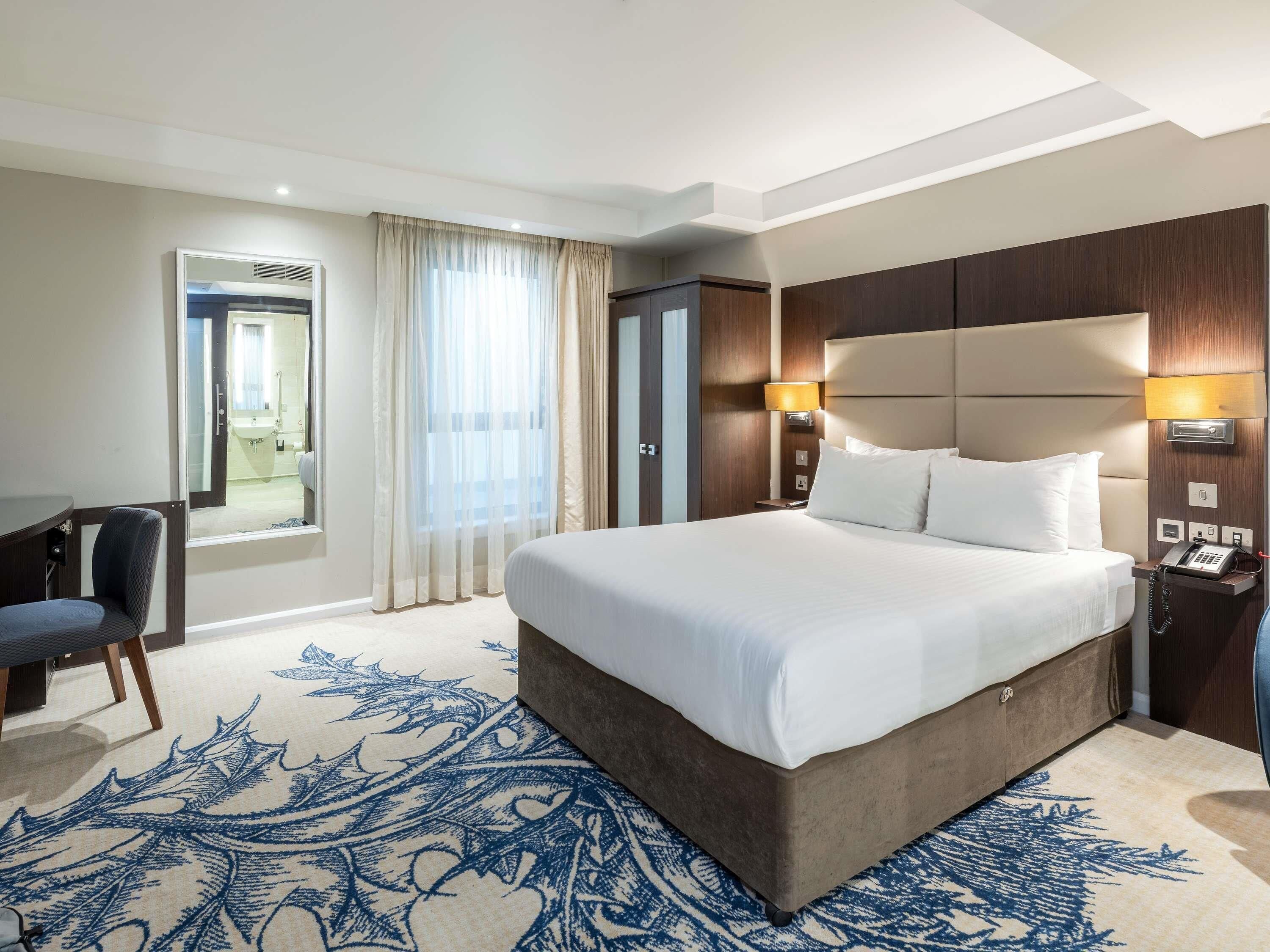 A hotel room with a large bed, a desk, a chair, a desk lamp, and a telephone.