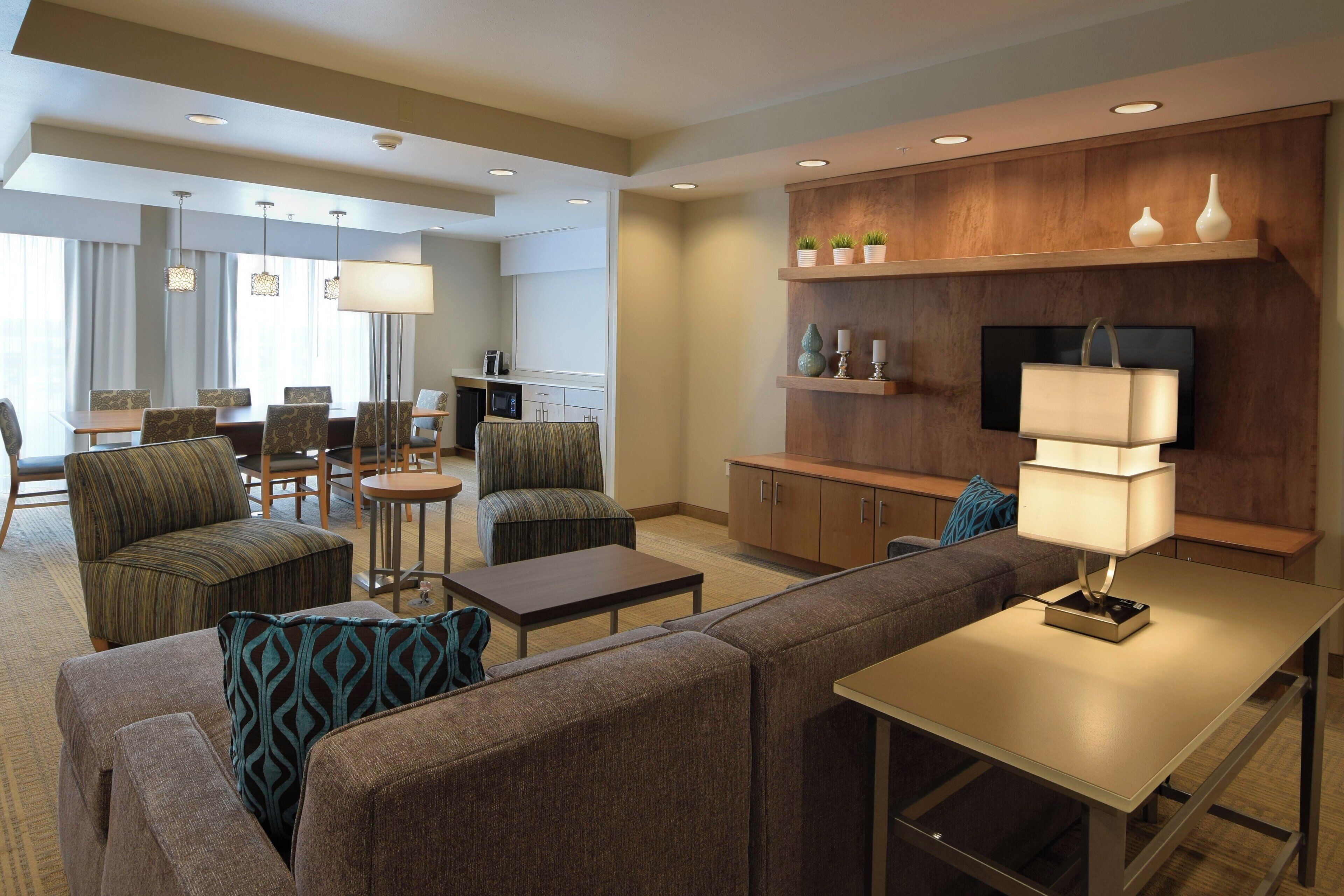 A hotel room with a sofa, armchair, dining table, and a TV mounted on a wooden panel.