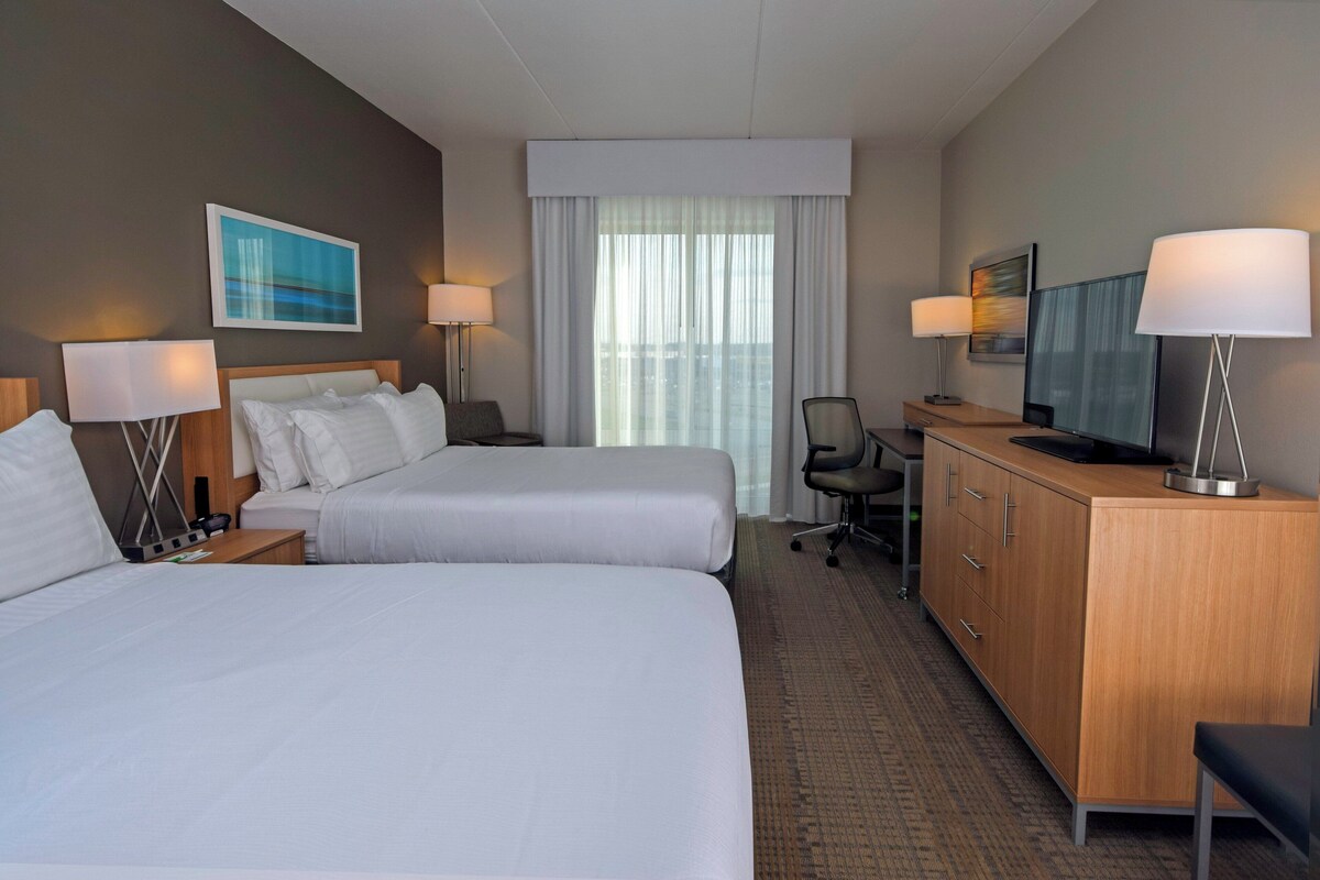 A hotel room with two beds, a desk, a television, and a large window with curtains.