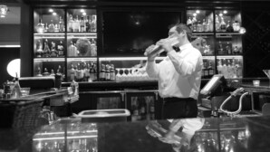 A bar with shelves of liquor bottles, a cash register, and a bartender preparing a drink. - Holiday Inn Cincinnati N - West Chester by IHG (West Chester)