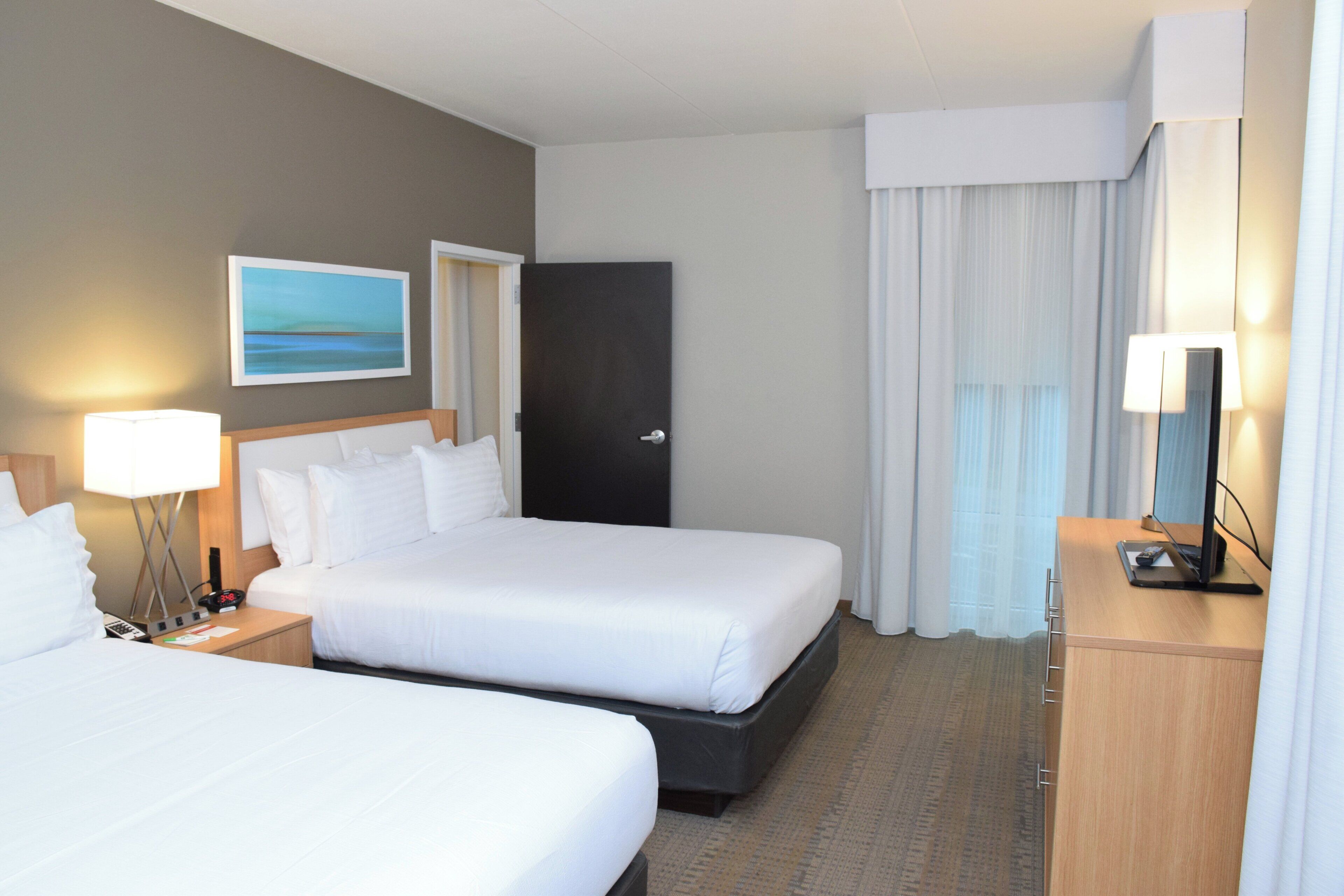 A hotel room with two beds, a desk, a lamp, a painting on the wall, and a door.