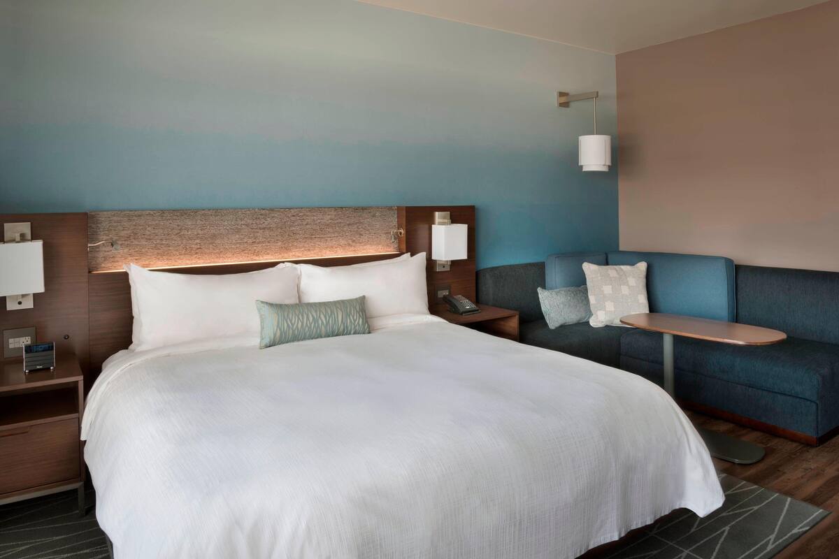 A hotel room with a large bed, a sofa, a nightstand with a lamp, and a wall-mounted phone.