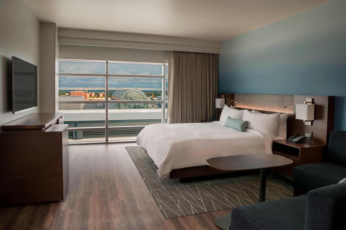 A hotel room with a large bed, a desk, a chair, a TV, and a view of a cityscape through a large window.