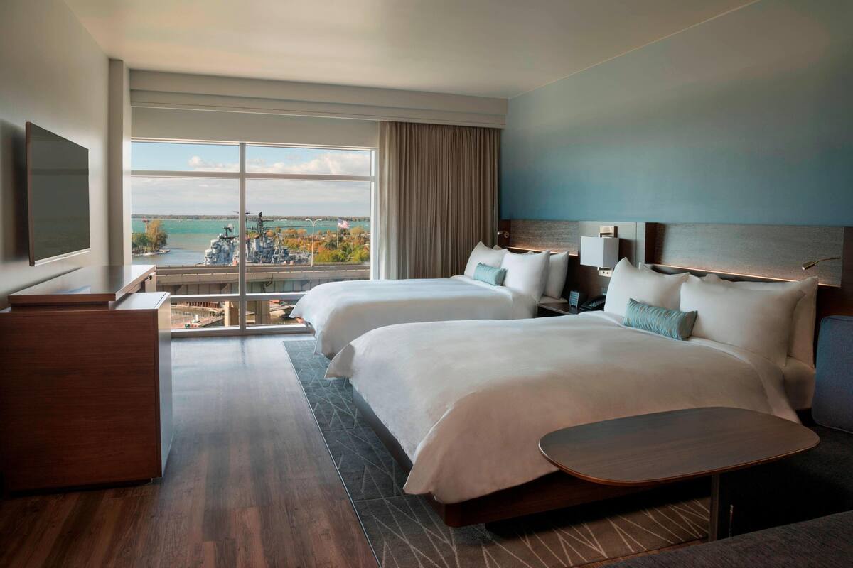 A hotel room with two beds, a desk, a TV, and a view of the city and water.