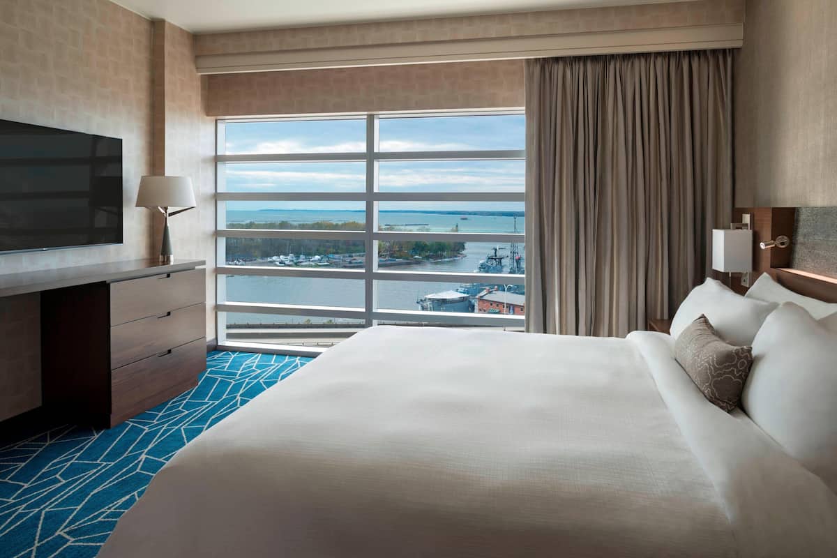 A hotel room with a large bed, a TV on a wooden dresser, and a view of a body of water and buildings.