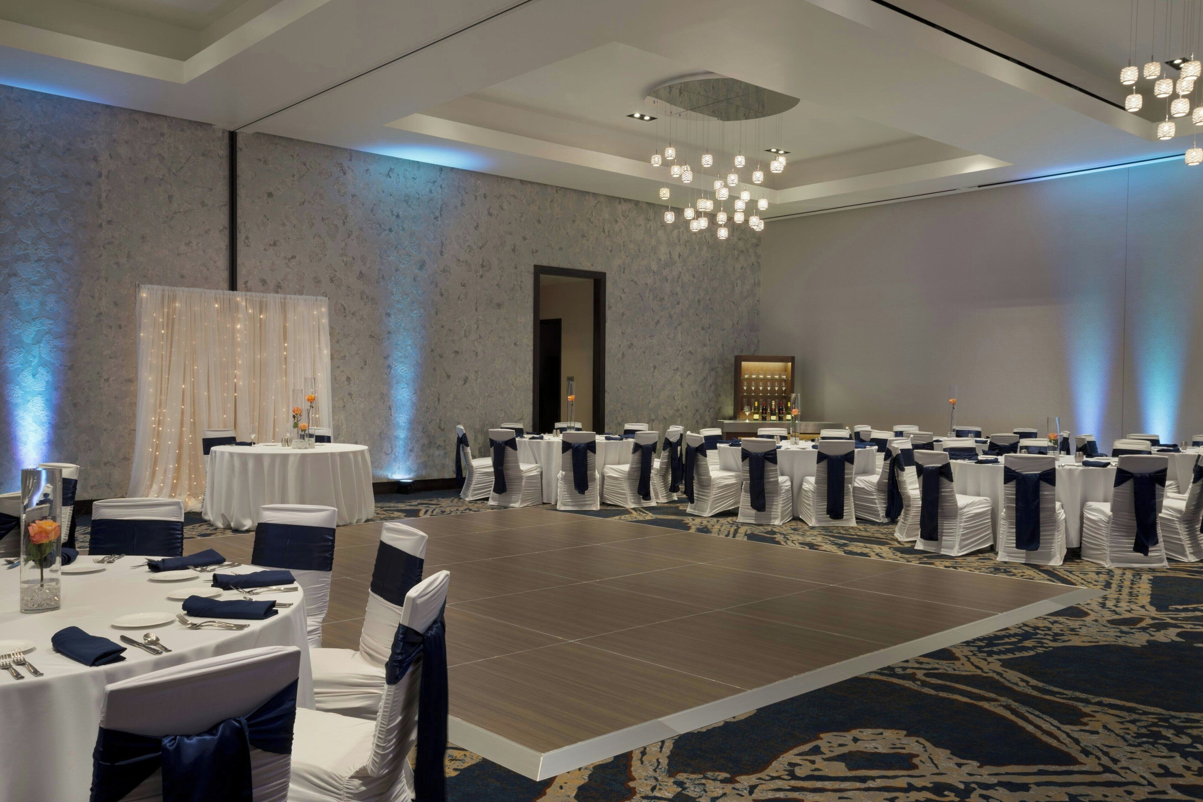 A conference room with round tables, chairs with blue sashes, a stage with a podium, and a chandelier.