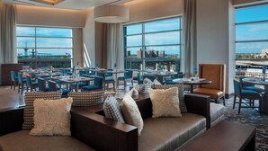 Un vestíbulo de hotel con zona de estar, mesas de comedor y grandes ventanales que ofrecen vistas del paisaje urbano.
