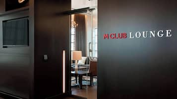 A modern lounge area with a glass partition, a sofa, a table, and a lamp. The text "M CLUB LOUNGE" is visible on the wall.
