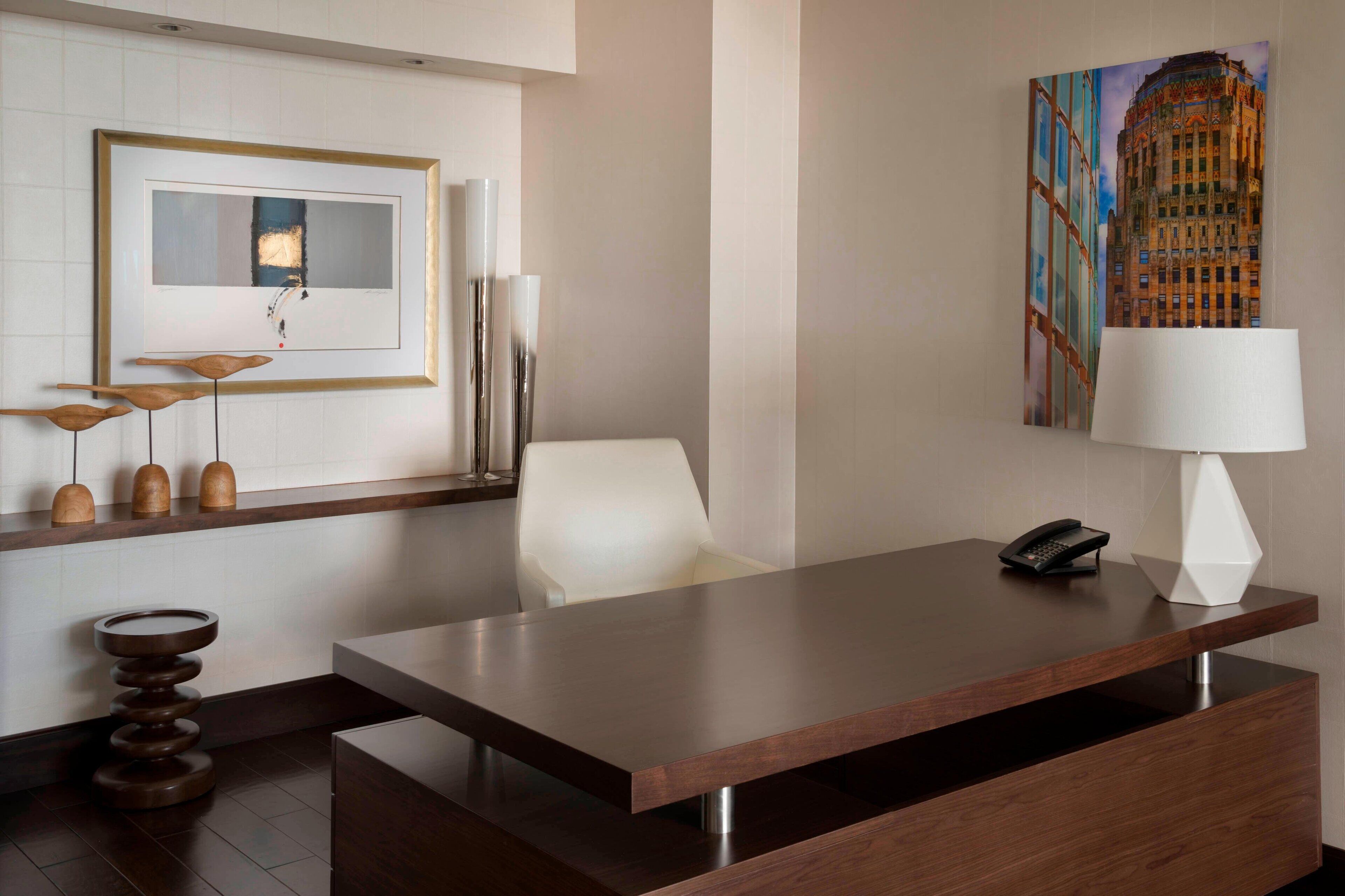 A modern hotel room with a wooden desk, a telephone, and a painting of a building on the wall.